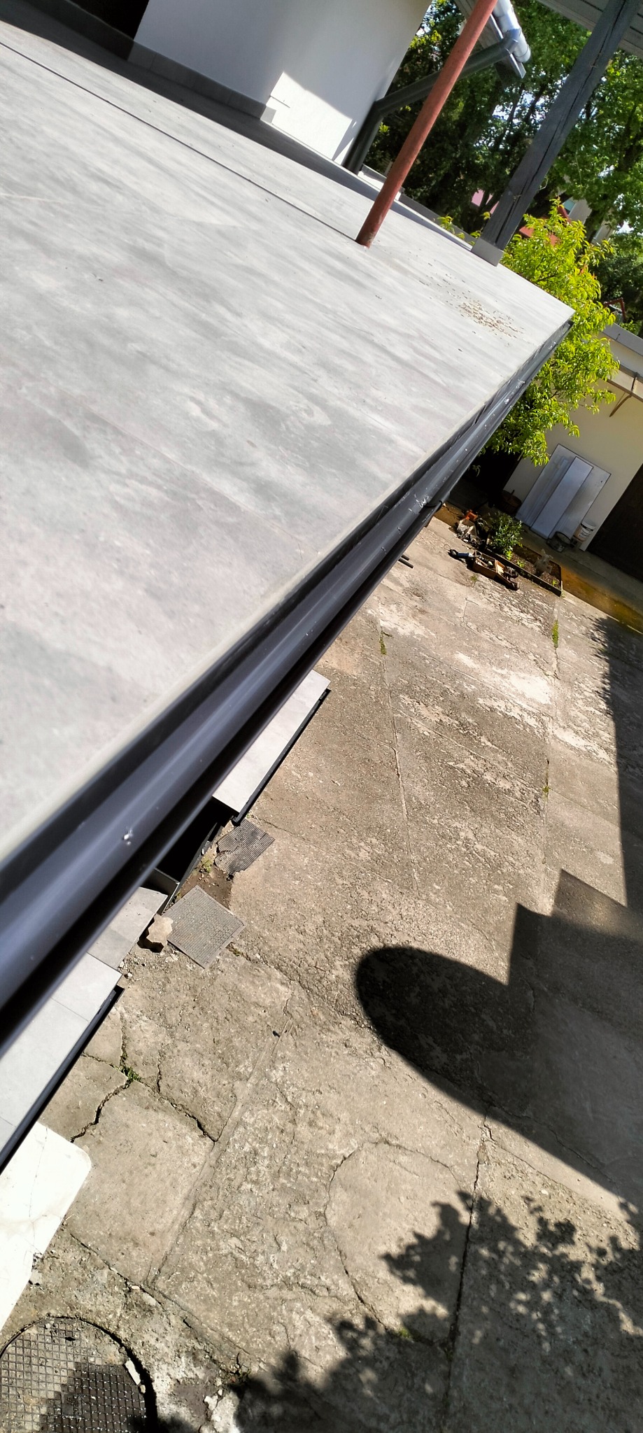 Wykończony taras z szarą, gładką powierzchnią i czarną listwą wykończeniową. Widoczne płytki i betonowa nawierzchnia. Ujęcie z góry, w słoneczny dzień.