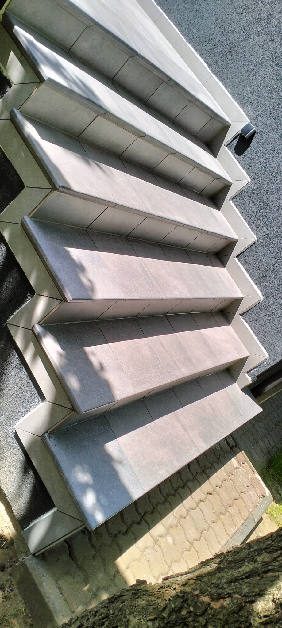 Kaskadowe schody zewnętrzne wyłożone szarą płytką, z widocznym fragmentem elewacji budynku o szarej strukturze i kostki brukowej. Ujęcie z dołu, w słoneczny dzień.