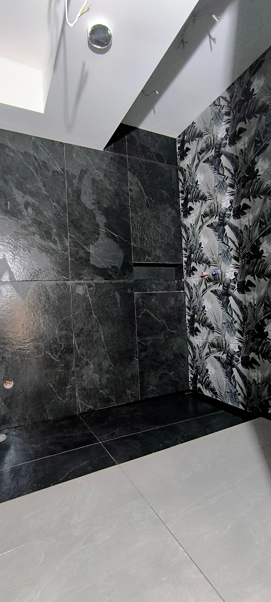 Częściowo wykończona łazienka z czarnymi płytkami imitującymi marmur na ścianach i podłodze, oraz ścianą z motywem roślinnym. Widoczne instalacje.