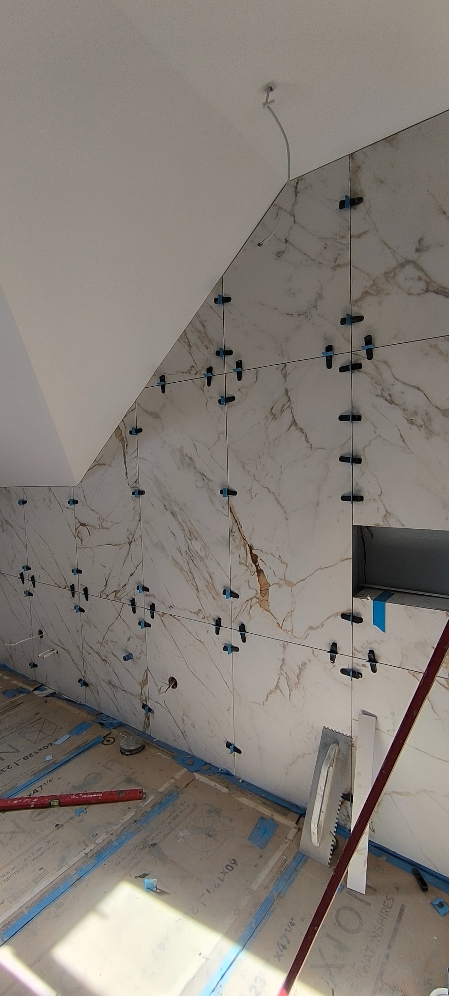 Ściana w trakcie układania płytek imitujących marmur, z użyciem klipsów poziomujących. Widoczne narzędzia glazurnicze i fragment podłogi zabezpieczonej kartonem.