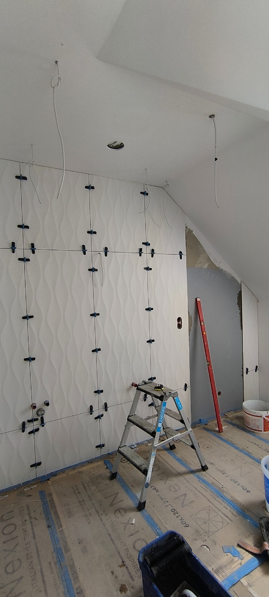 Ściana z falistych, białych płytek w trakcie montażu. Krzyżyki dystansowe, drabina Zarges, poziomica. Widoczne kable oświetleniowe. Podłoga zabezpieczona kartonem.