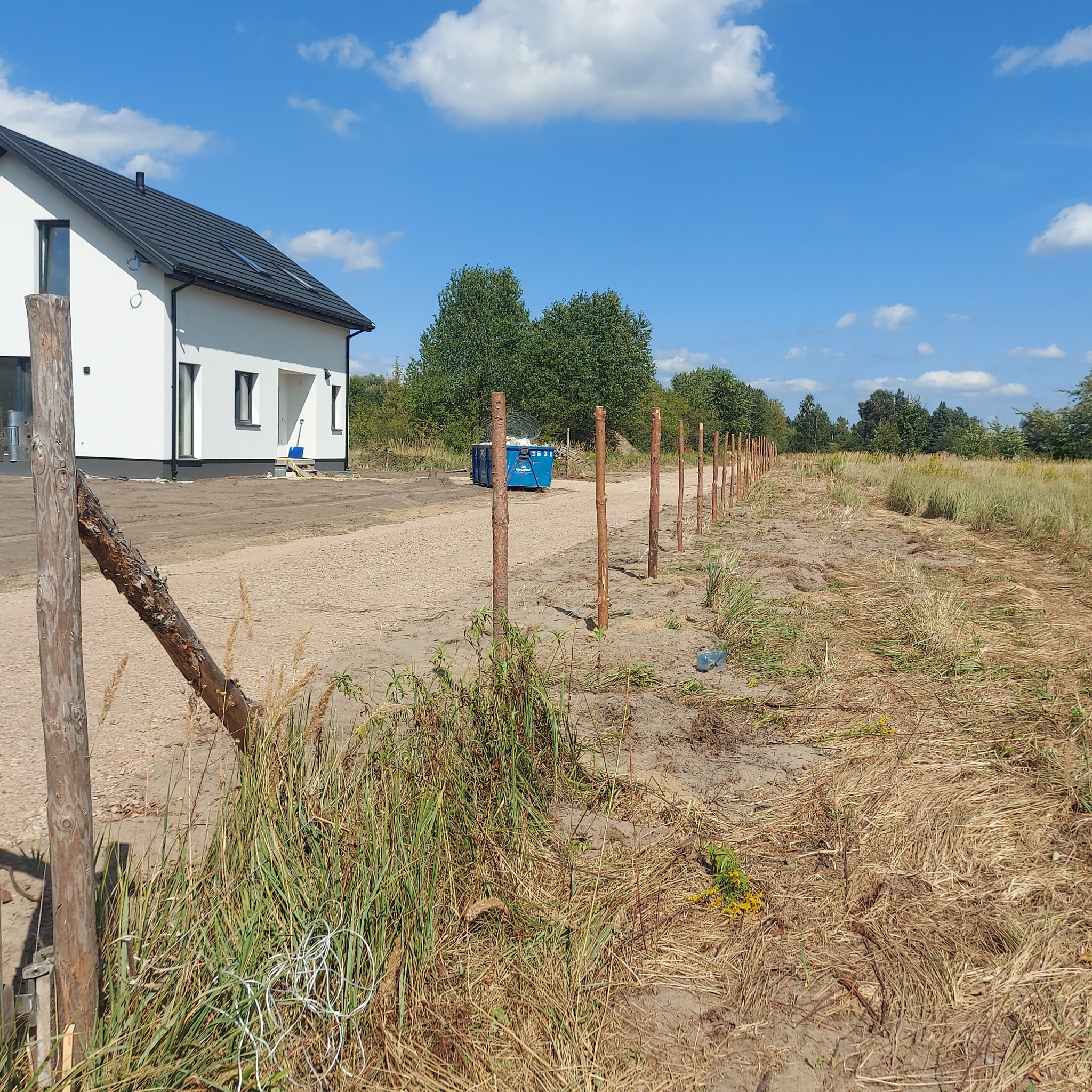 Wkopane słupki ogrodzeniowe w ziemi, widoczne na tle domu z białymi ścianami i czarnym dachem, błękitne niebo. Etap początkowy montażu ogrodzenia.