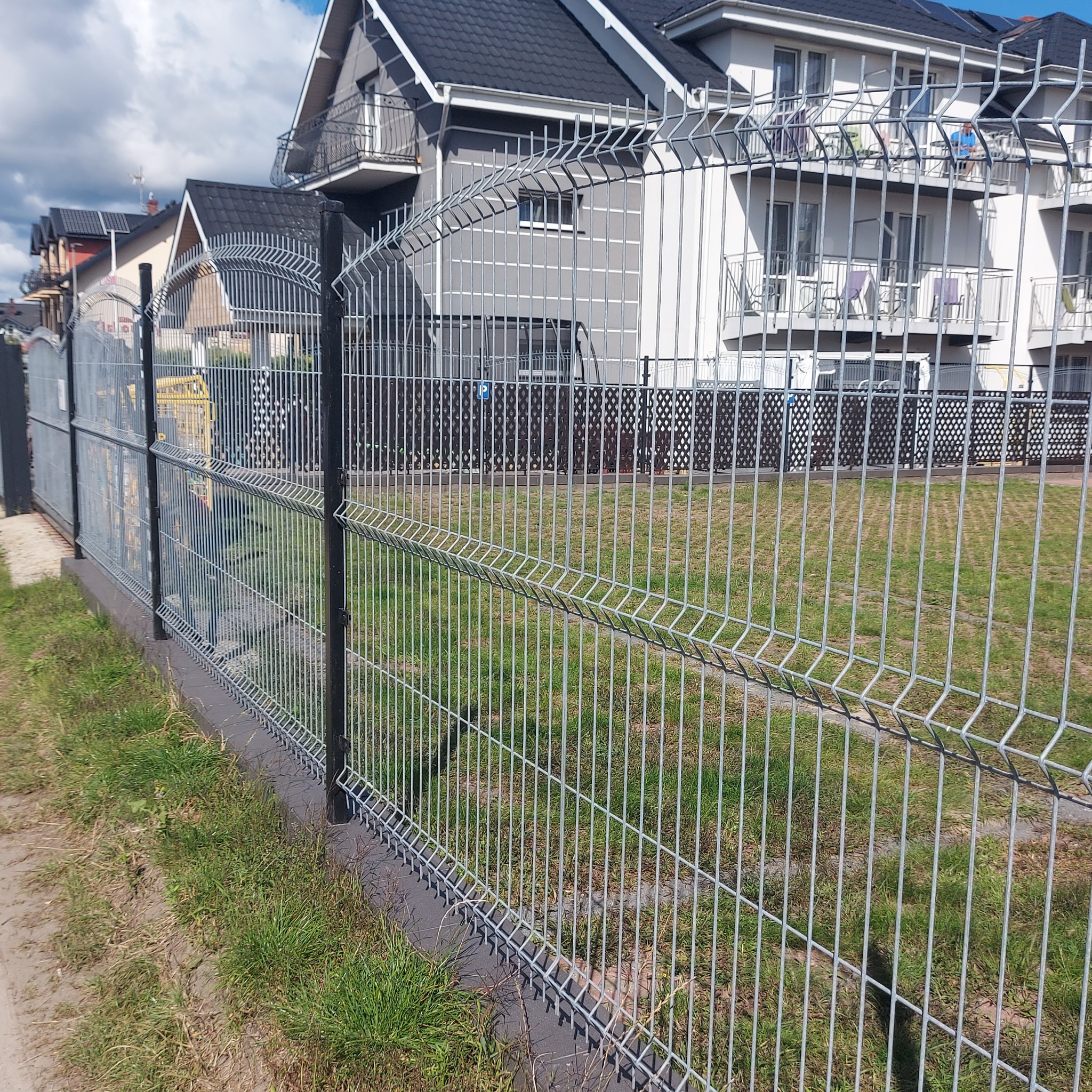 Nowoczesne ogrodzenie panelowe z metalu, z czarnymi słupkami, na tle domów jednorodzinnych. Widok z poziomu gruntu, zieleń trawy.