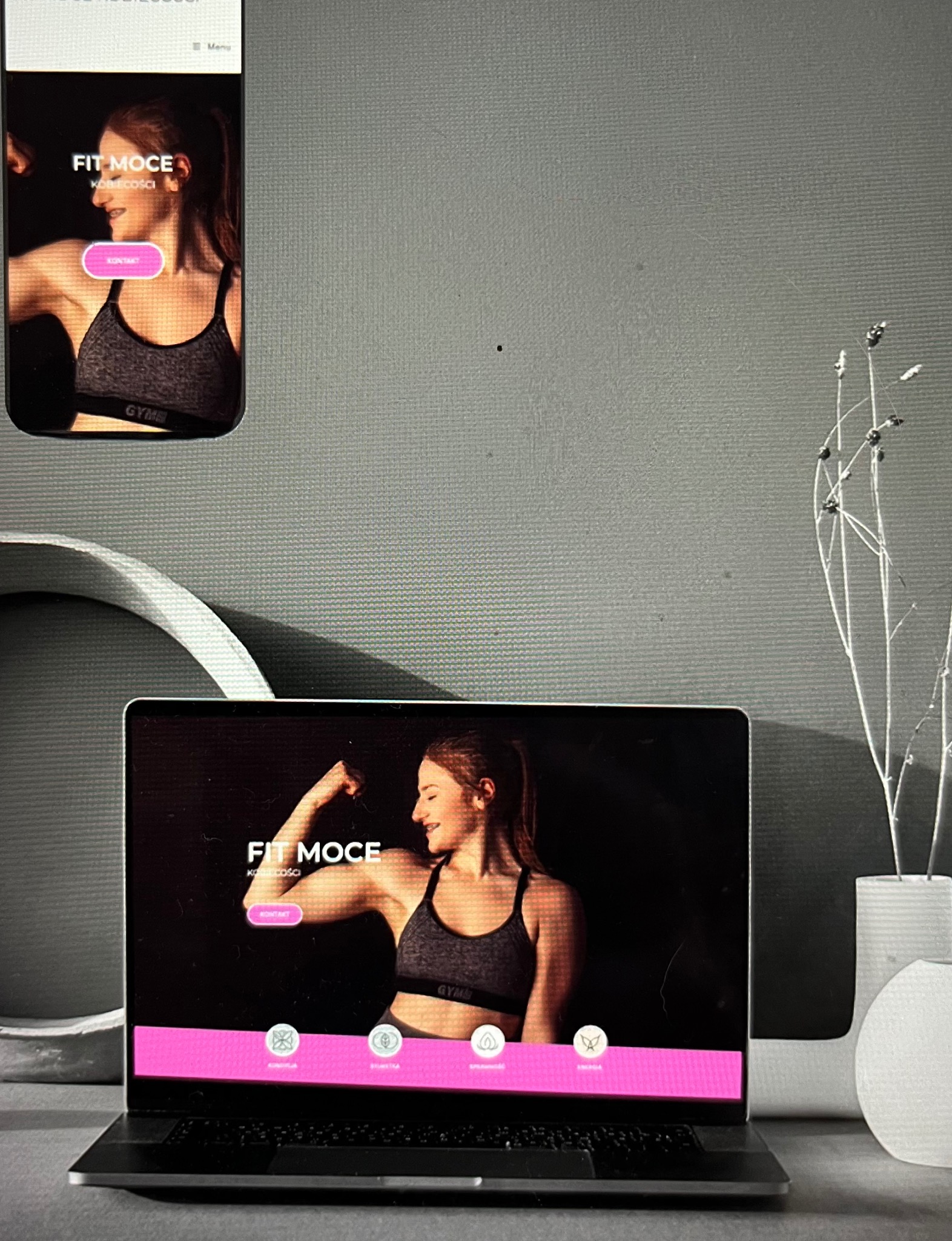 Ekran laptopa i smartfona wyświetlające stronę internetową z wizerunkiem kobiety prezentującej muskulaturę, obok wazon z suchymi gałązkami na tle szarej ściany.