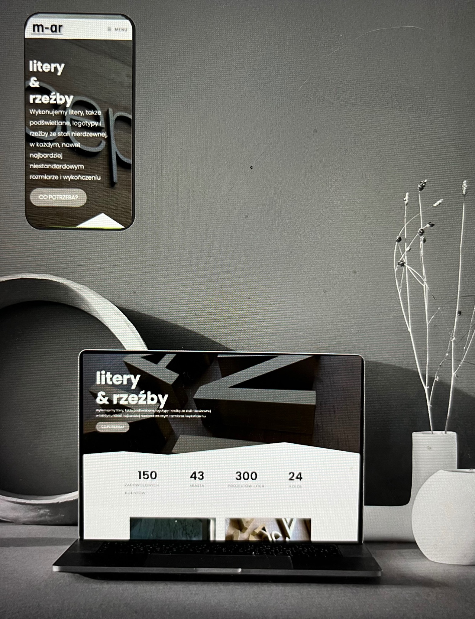 Ekran laptopa i smartfona prezentujące stronę internetową firmy zajmującej się literami i rzeźbami, obok dekoracyjny wazon z suszem, stonowana kolorystyka.
