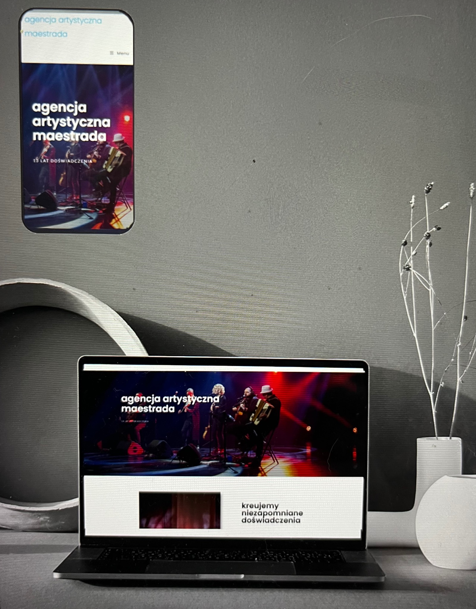Prezentacja strony internetowej agencji artystycznej 'Maestrada' na laptopie i smartfonie, z zespołem muzycznym na scenie w tle, minimalistyczna dekoracja z białych wazonów i gałązek.
