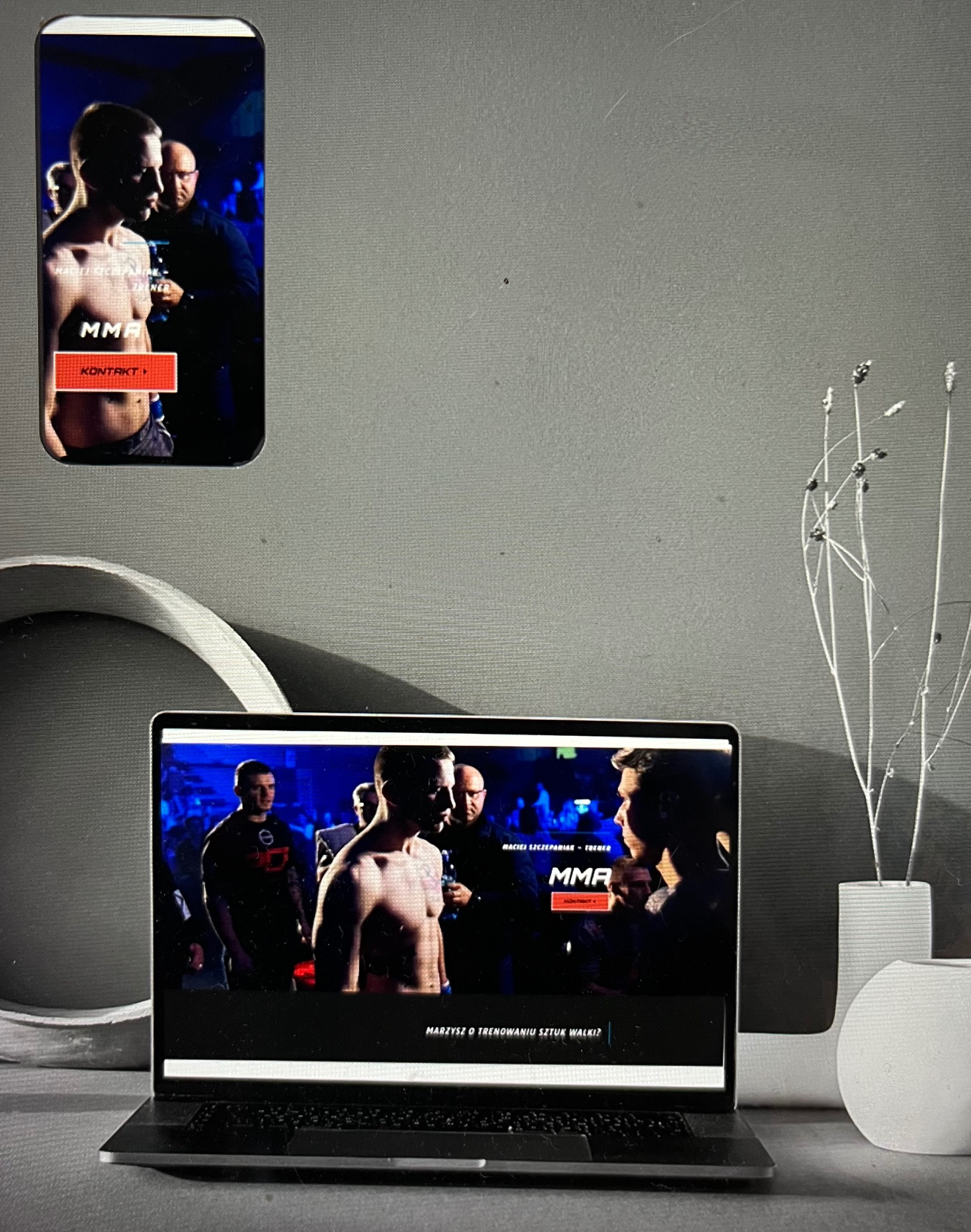 Ekran laptopa i smartfona wyświetlające tę samą stronę internetową z zawodnikami MMA przed walką, minimalistyczne tło z dekoracyjnym wazonem.