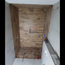 Usługi wykończeniowe - Montaż płytek imitujących drewno na ścianie w łazience, widoczne krzyżyki dystansowe, poziomica i przewody pomiarowe.