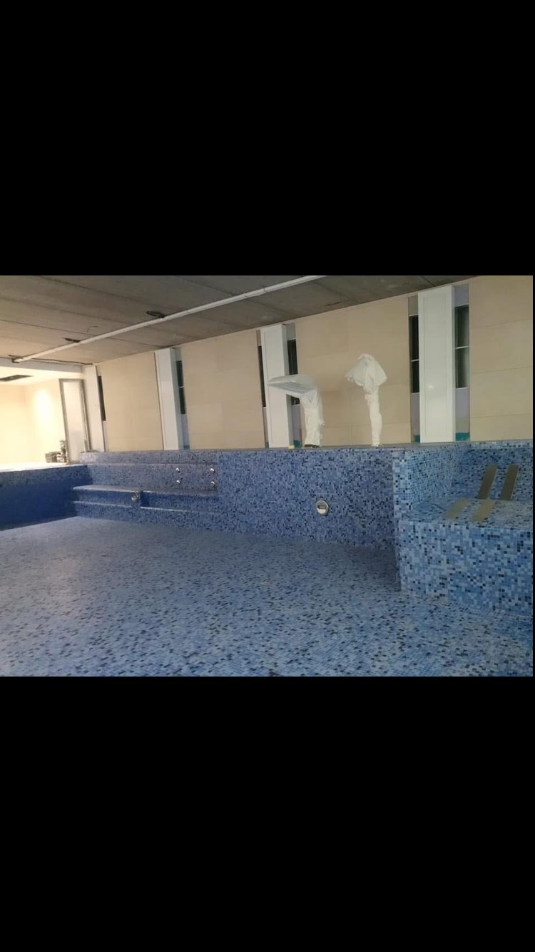 Wnętrze basenu w trakcie wykańczania, wyłożonego niebieską mozaiką, z widocznymi schodkami i elementami oświetlenia w ścianach, zabezpieczone elementy pomieszczenia w tle.