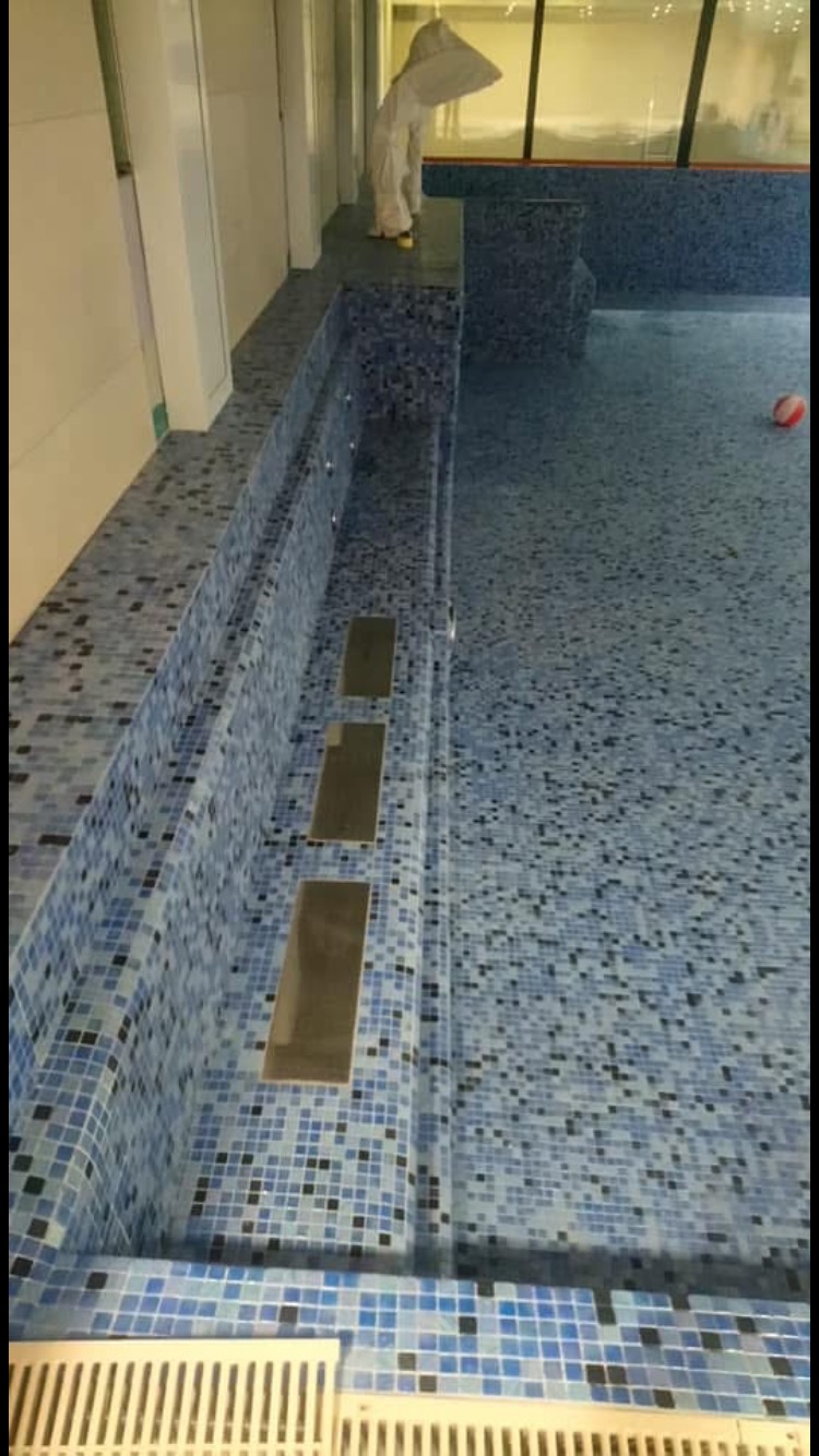 Niebieska mozaika w basenie z metalowymi wnękami w ścianie, osoba w kombinezonie ochronnym stoi na brzegu, widoczna kratka odpływowa i piłka w basenie.