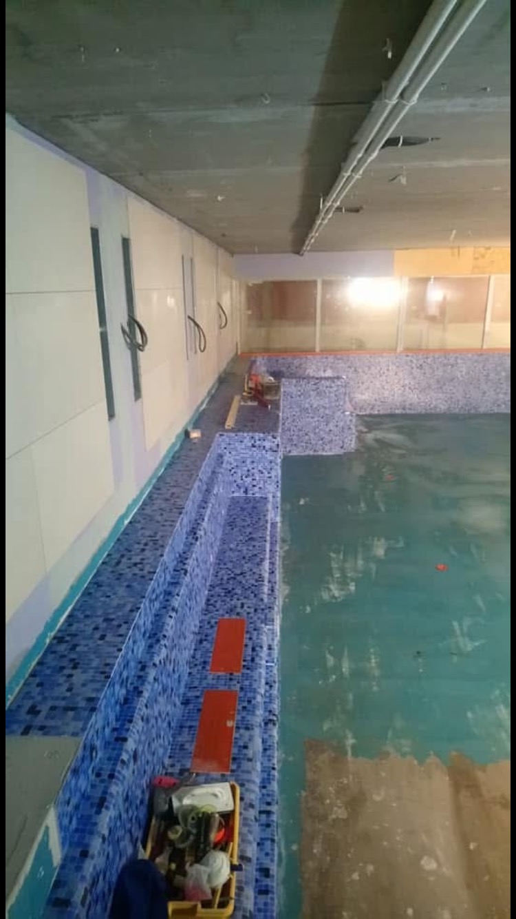 Wnętrze basenu w trakcie wykańczania, widoczne niebieskie mozaiki na ścianach i stopniach, żółty pojemnik z narzędziami oraz fragmenty podłogi bez wykończenia.