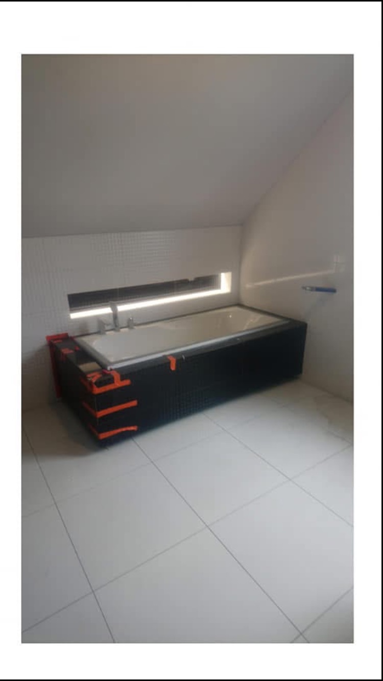 Wanna obudowana czarnym panelem z pomarańczowymi akcentami, zabezpieczona folią ochronną, w łazience z białymi płytkami i oświetleniem LED w niszy ściennej.