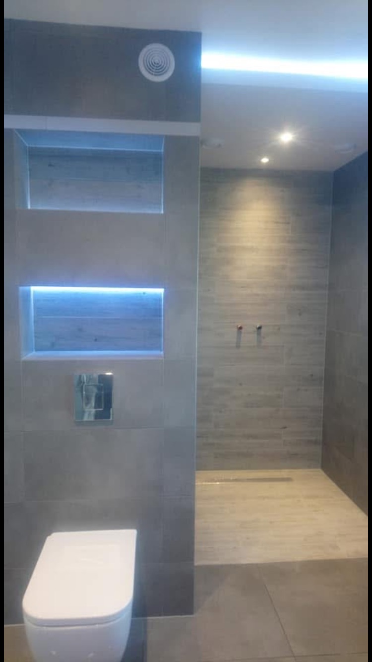 Nowoczesna łazienka z szarymi płytkami, podświetlanymi wnękami i toaletą wiszącą, widoczny odpływ liniowy w strefie prysznicowej.