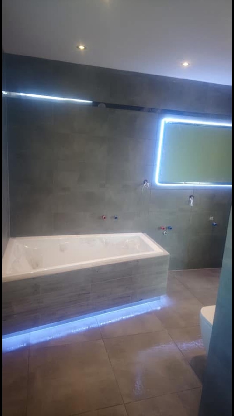 Nowoczesna łazienka z podświetlaną wanną i oknem, szare płytki na ścianach i podłodze, minimalistyczny design, widoczne instalacje wodne przed montażem armatury.