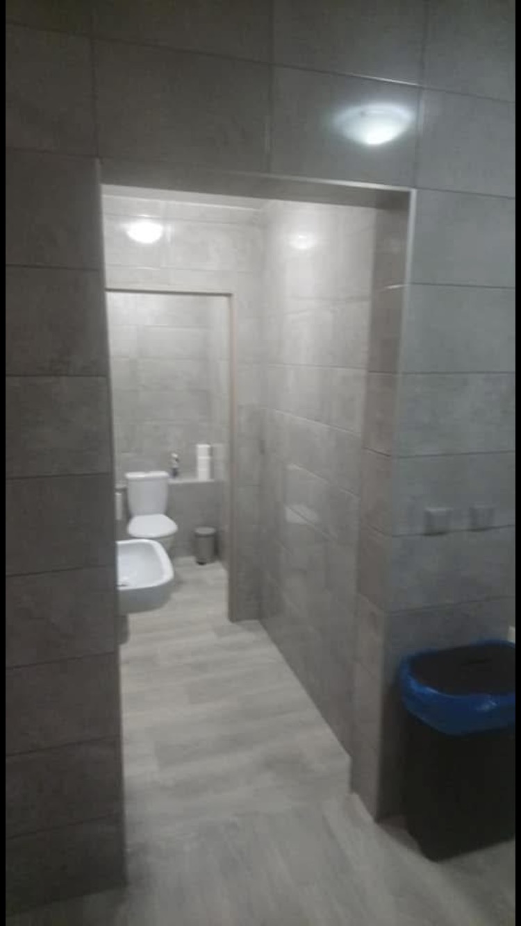 Szara łazienka z toaletą, bidetem i koszem na śmieci, wykończona płytkami w odcieniach szarości i panelami podłogowymi, widoczna przez wąskie przejście.