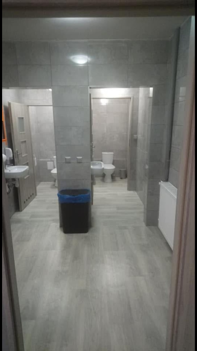 Wnętrze publicznej toalety z szarymi płytkami, umywalkami i toaletami za czarnym koszem na śmieci, widok z perspektywy wejścia.