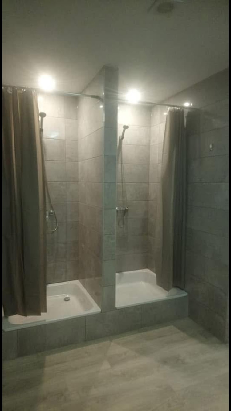 Dwie kabiny prysznicowe oddzielone ścianką, wykończone szarą glazurą i zasłonami w kolorze beżowym, z białymi brodzikami i drewnopodobną podłogą.