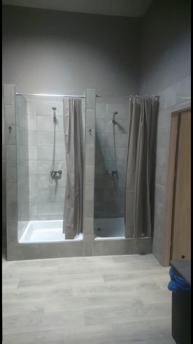 Dwie kabiny prysznicowe z szarymi płytkami i zasłonami, białe brodziki, jasna podłoga imitująca drewno, szare ściany.