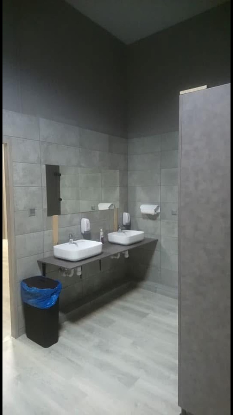 Szara łazienka publiczna z dwoma umywalkami nablatowymi, dozownikami mydła i ręcznikami papierowymi, wykończona płytkami ceramicznymi w odcieniach szarości i panelami podłogowymi imitującymi drewno...