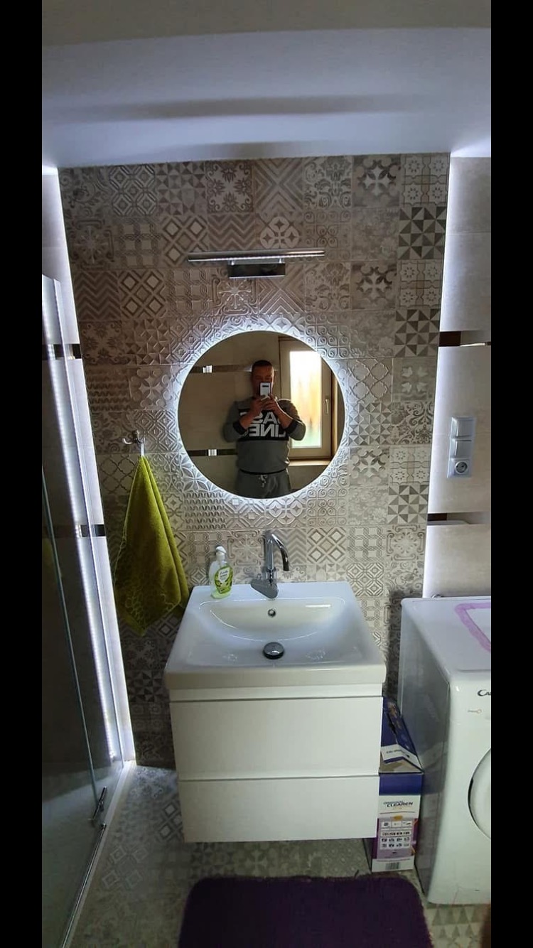 Wykończona łazienka z umywalką nablatową, okrągłym lustrem z podświetleniem LED i wzorzystymi płytkami na ścianie, widoczny fragment pralki i odbicie osoby robiącej zdjęcie.