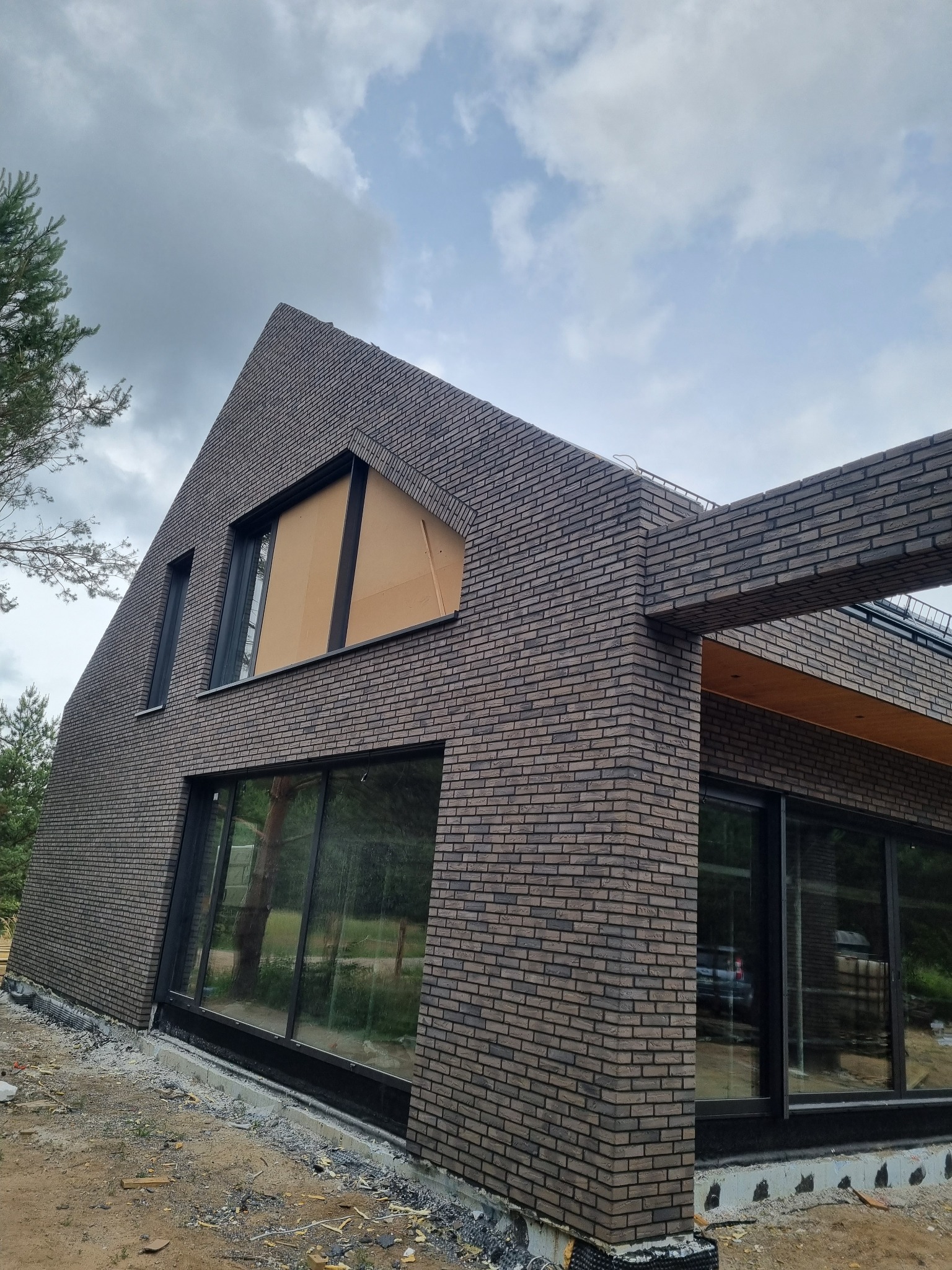 Nowoczesny dom z elewacją z brązowej cegły klinkierowej i dużymi oknami, częściowo zakrytymi kartonem, widok z dołu na tle pochmurnego nieba.