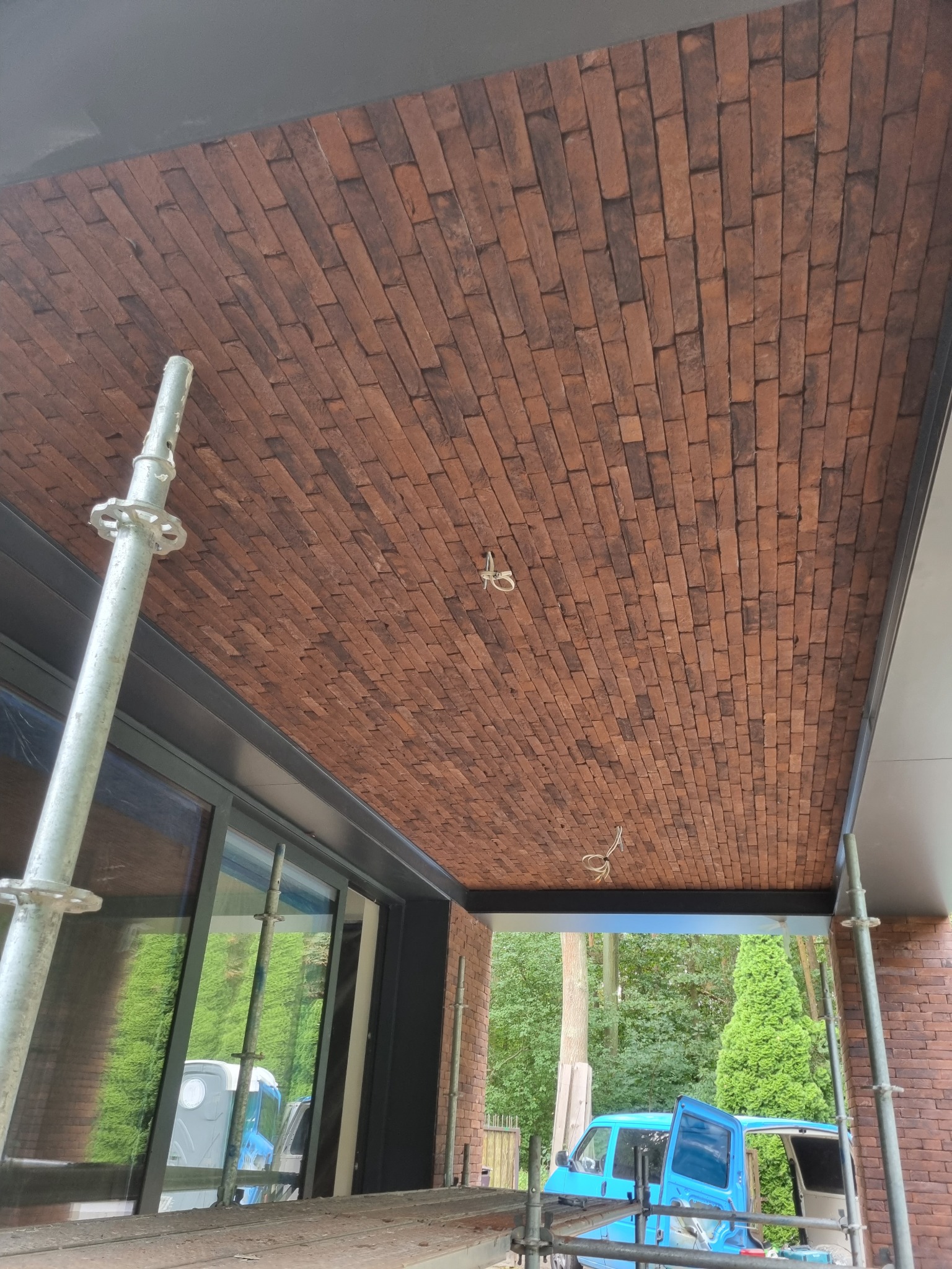 Ceglana podbitka dachu na zewnątrz budynku, widok z rusztowania, z widocznym fragmentem okna i niebieskim samochodem w tle.