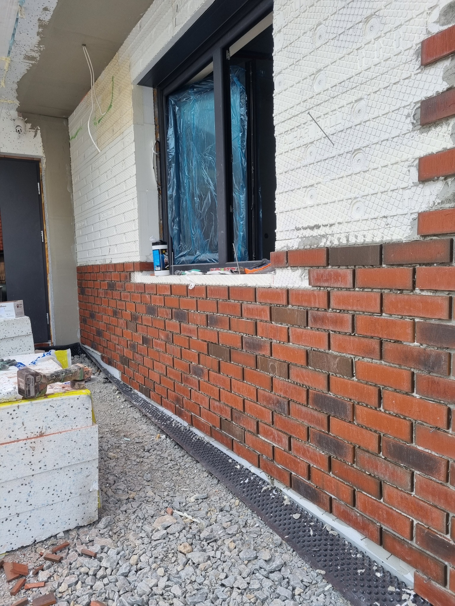 Ceglana okładzina elewacyjna w trakcie montażu, widoczne warstwy izolacji i folia ochronna na oknie, narzędzia i materiały budowlane w pobliżu.