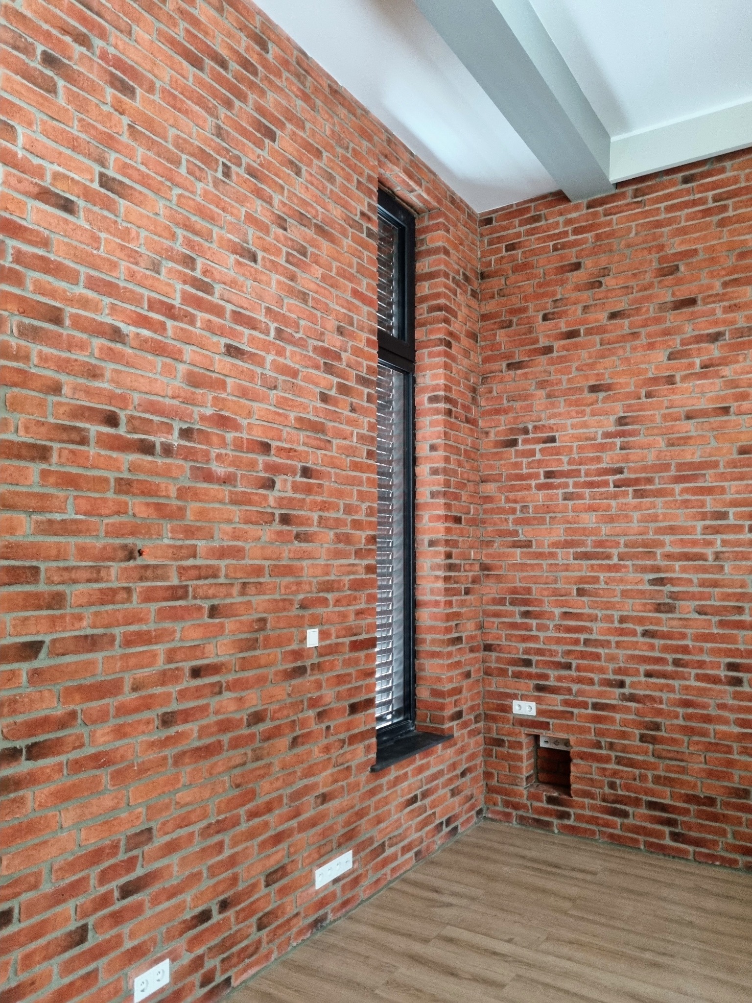 Wnętrze z ceglaną ścianą, wąskie okno z żaluzjami, drewniana podłoga i biały sufit z szarymi belkami.