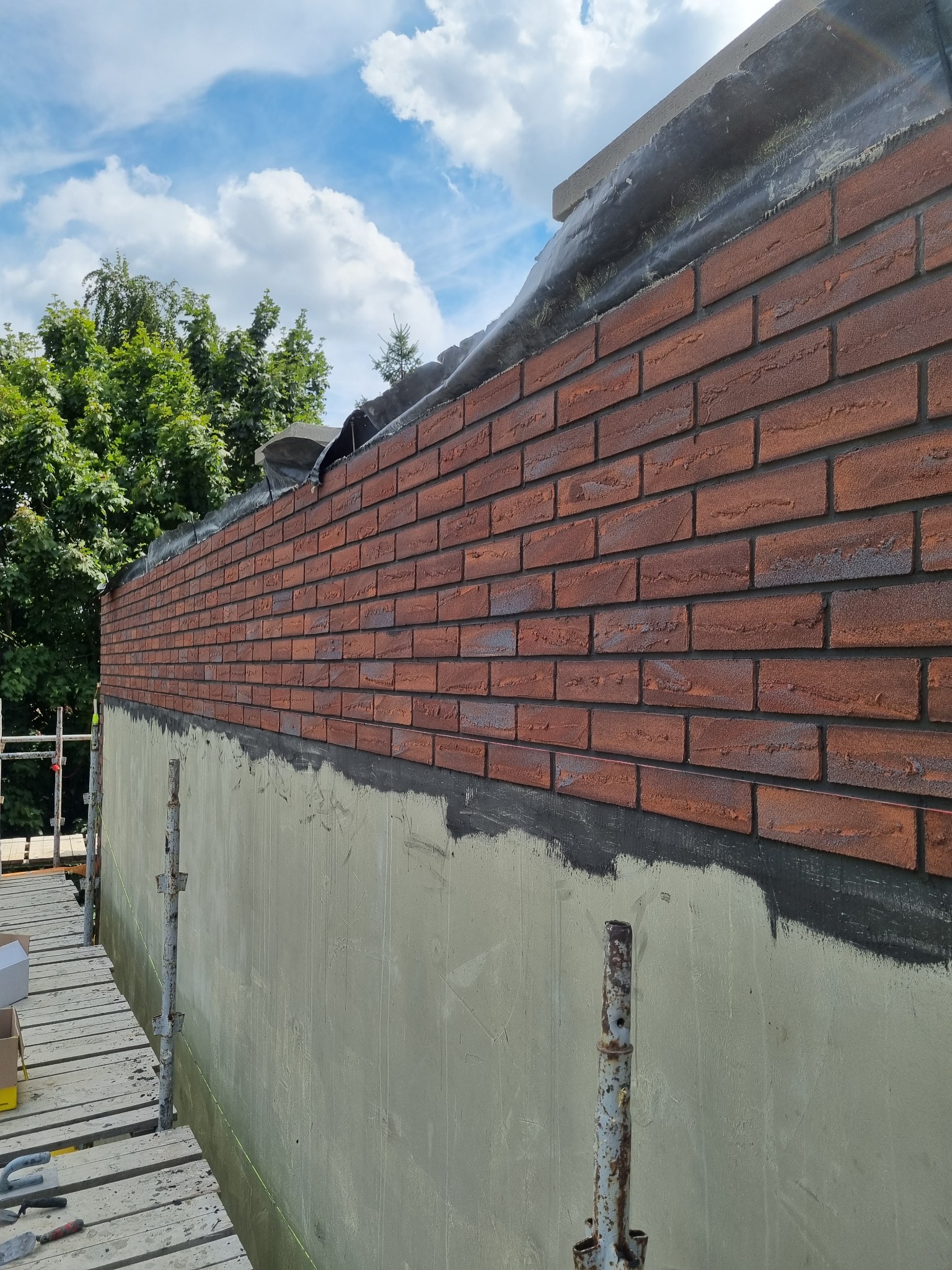 Częściowo wykończona elewacja budynku z cegły klinkierowej, widoczna z rusztowania, z zielenią drzew i błękitnym niebem w tle.