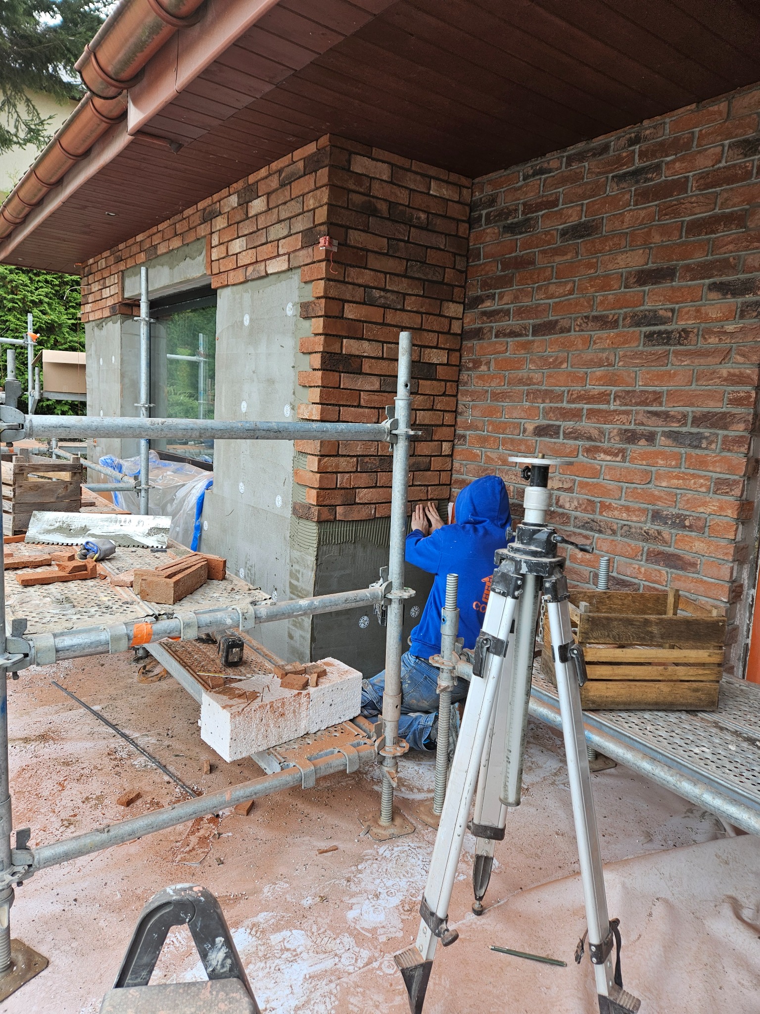 Pracownik w niebieskiej bluzie na rusztowaniu układa cegły klinkierowe na elewacji budynku, widoczny poziomica na trójnogu i skrzynki z cegłami.