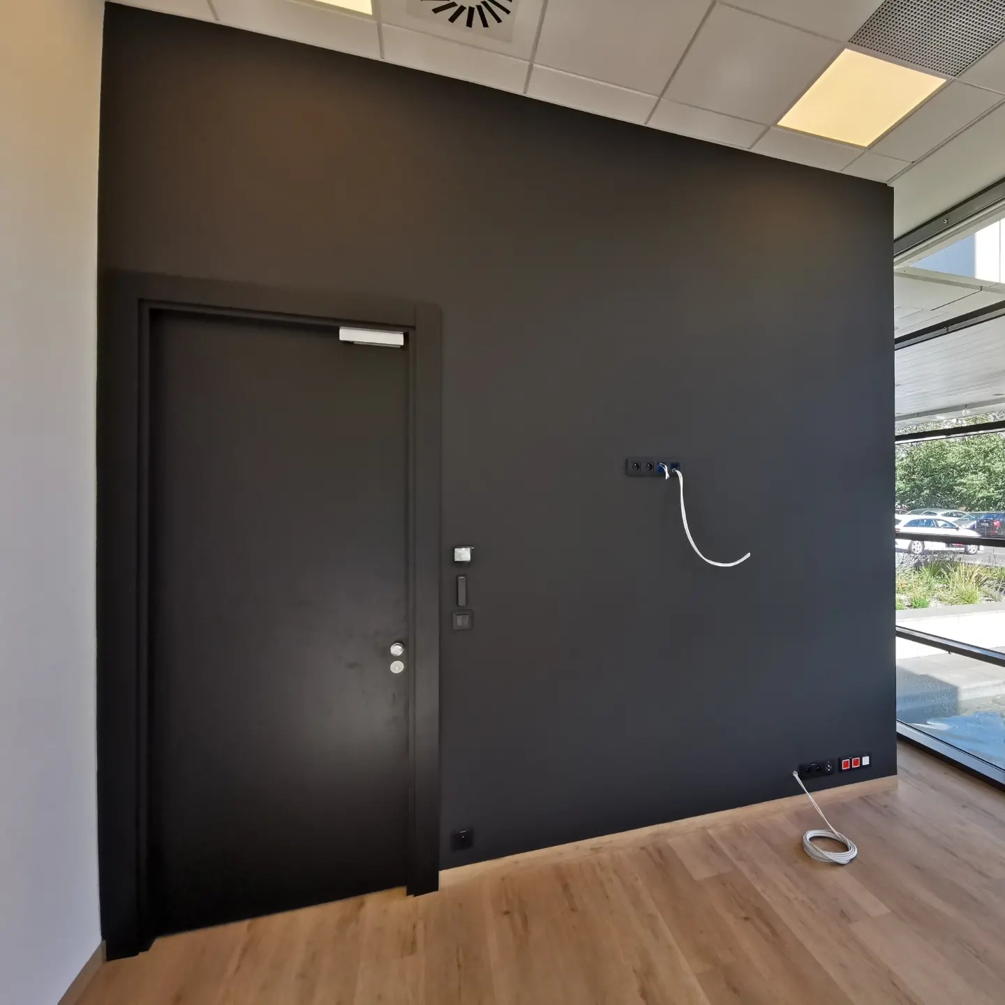 Ciemnoszara ściana w biurze po malowaniu, widoczna czarna, zamknięta brama przeciwpożarowa, gniazdka elektryczne i kabel wystający ze ściany.