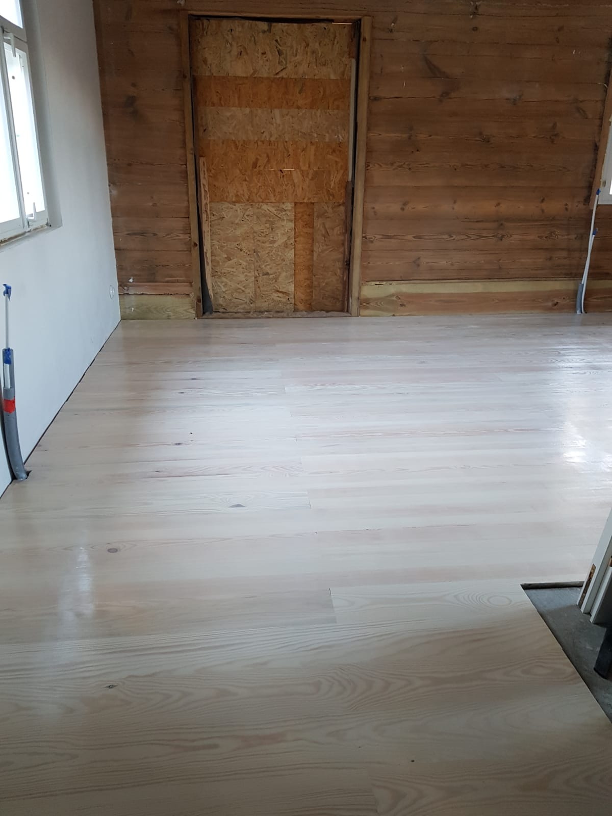 Jasna, odnowiona podłoga drewniana w pomieszczeniu z drewnianą ścianą i zamurowanym otworem drzwiowym.