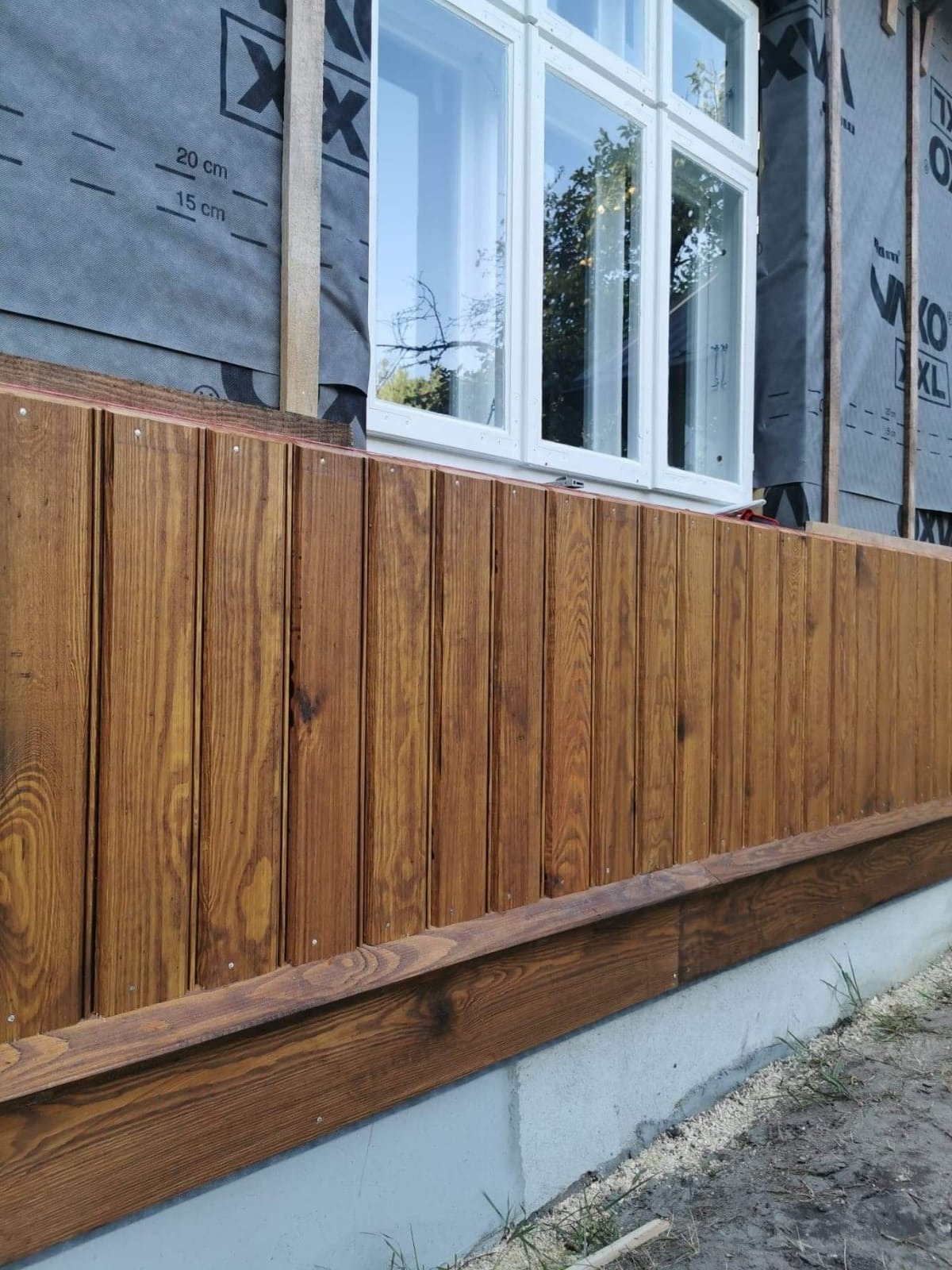 Fragment elewacji budynku z drewnianą boazerią w kolorze brązowym, widoczna izolacja przeciwwilgociowa i białe okno.