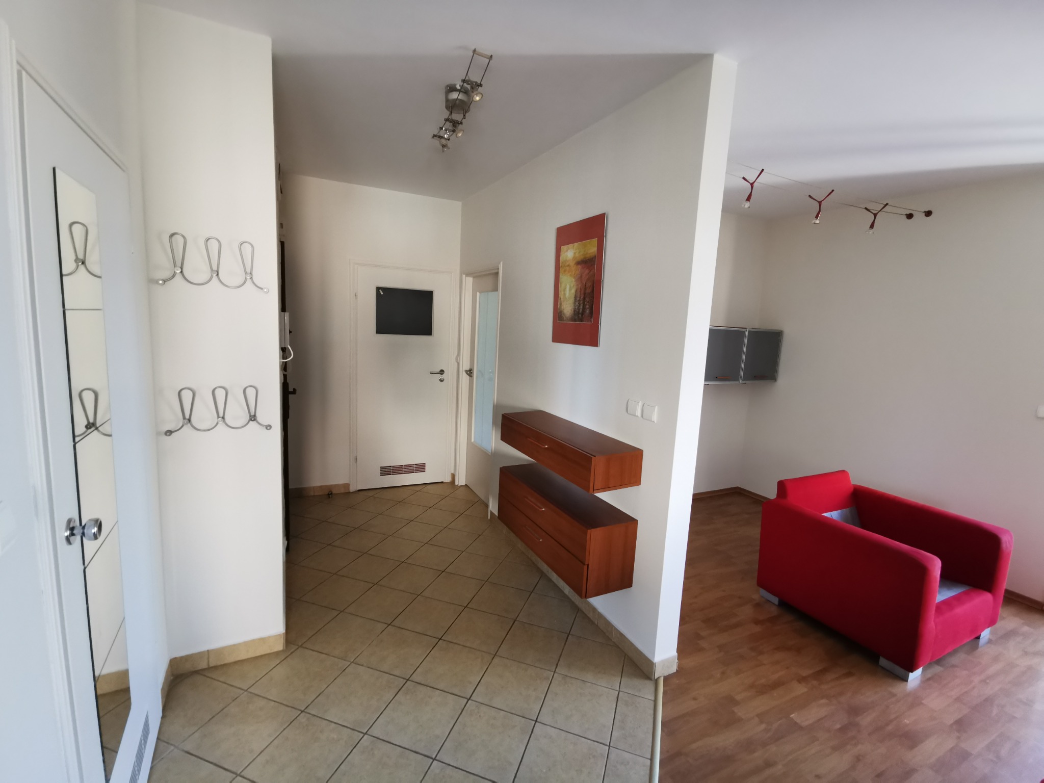 Jasne wnętrze mieszkania z przedpokojem i salonem, widoczne wieszaki na ścianie, dwie wiszące komody z szufladami, czerwony fotel i lampy sufitowe.