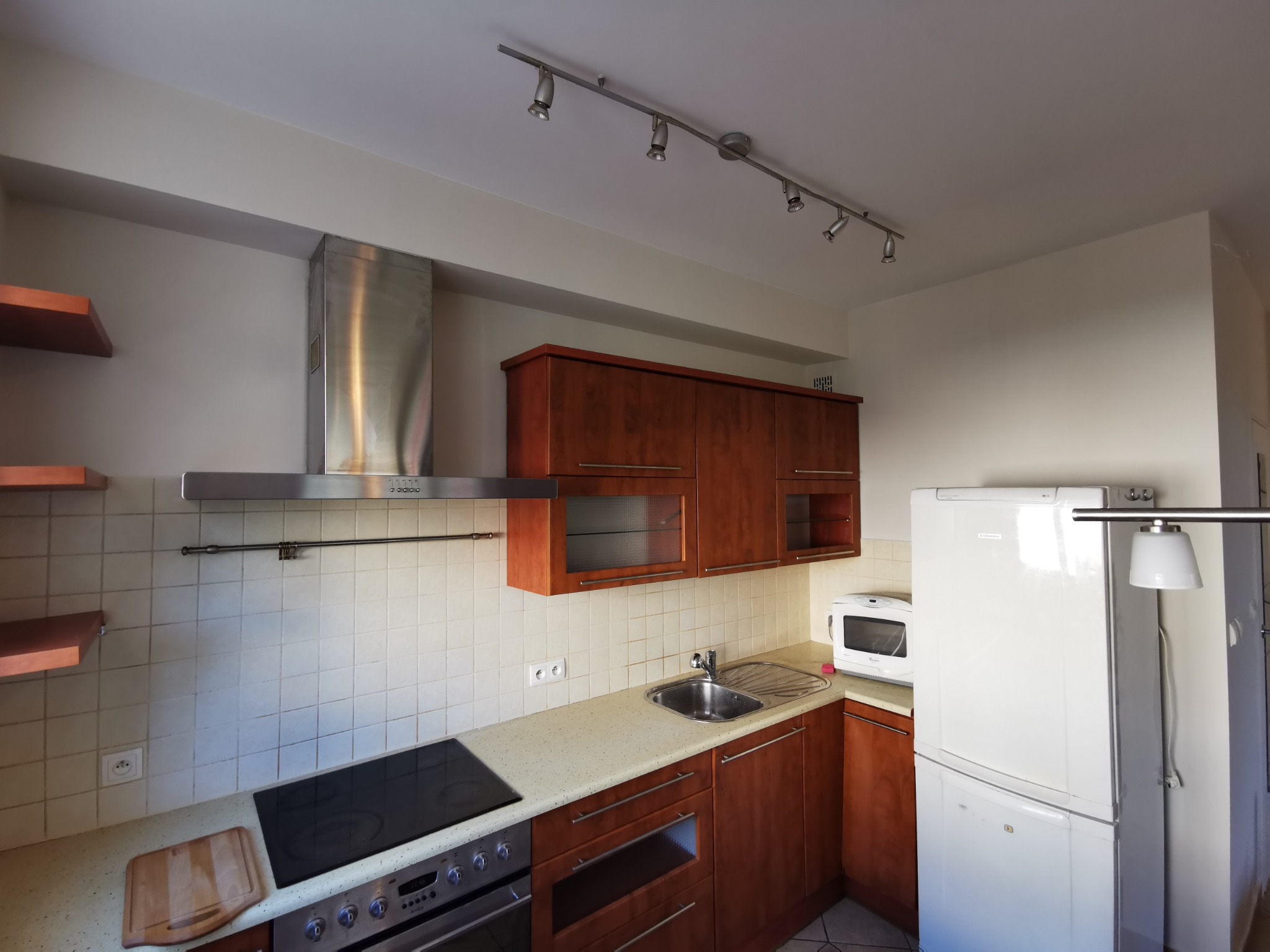 Kuchnia z drewnianymi szafkami, białymi ścianami i jasnym blatem, oświetlona listwą z reflektorami.