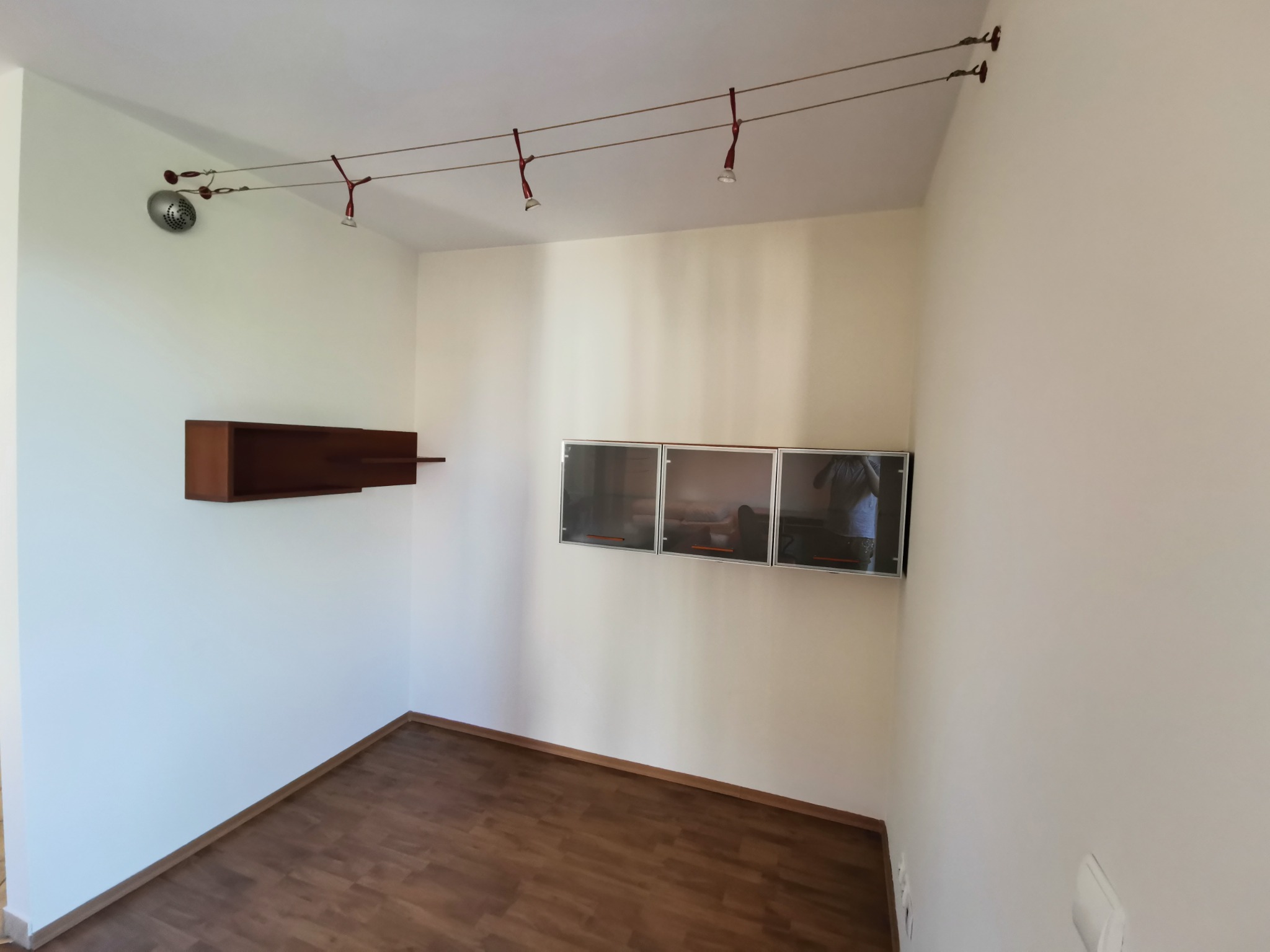 Pomalowane na biało wnętrze pokoju z drewnianą podłogą, brązową półką i trzema wiszącymi szafkami z szarymi frontami, oświetlone systemem szynowym z czerwonymi reflektorami.