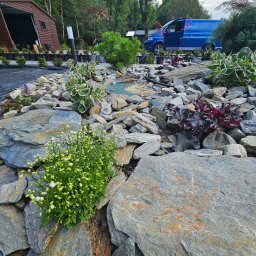 Green Dream Natalia Malczyk - Aranżacja skalna w ogrodzie z kwitnącymi białymi kwiatami na pierwszym planie, różnorodne rośliny ozdobne, niebieski samochód dostawczy w tle, widoczne podłoże wyłożone agrowłókniną.