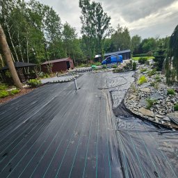 Green Dream Natalia Malczyk - Rozłożona czarna agrotkanina z zielonymi paskami na przygotowanym terenie pod ogród, widoczne elementy przyszłego skalniaka z kamieniami, w tle altana, budynek gospodarczy i niebieski samochód...