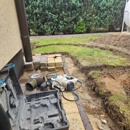 Green Dream Natalia Malczyk - Przygotowanie terenu pod instalację nawadniania: wykopane rowki w ziemi obok ułożonej kostki, narzędzia pracy - wiertarka udarowa i otwarty kufer narzędziowy leżą na żółtej kostce brukowej, w tle...