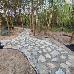 Green Dream Natalia Malczyk - Ścieżka z nieregularnych, jasnych kamieni, świeżo ułożonych w szarym cemencie, wijąca się przez las brzozowo-sosnowy, z widocznymi elementami instalacji ogrodowych w tle.