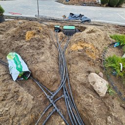 Green Dream Natalia Malczyk - Rozbudowany system nawadniania w trakcie instalacji, widoczne czarne rury i złączki w wykopie, obok worek ziemi ogrodniczej i kamień, w tle ułożona kostka brukowa i zaparkowany niebieski bus.