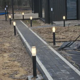 Ścieżka z ciemnej kostki brukowej obramowana jasnym kamieniem, oświetlona szeregiem nowoczesnych lamp ogrodowych, prowadząca do drewnianego budynku w tle.