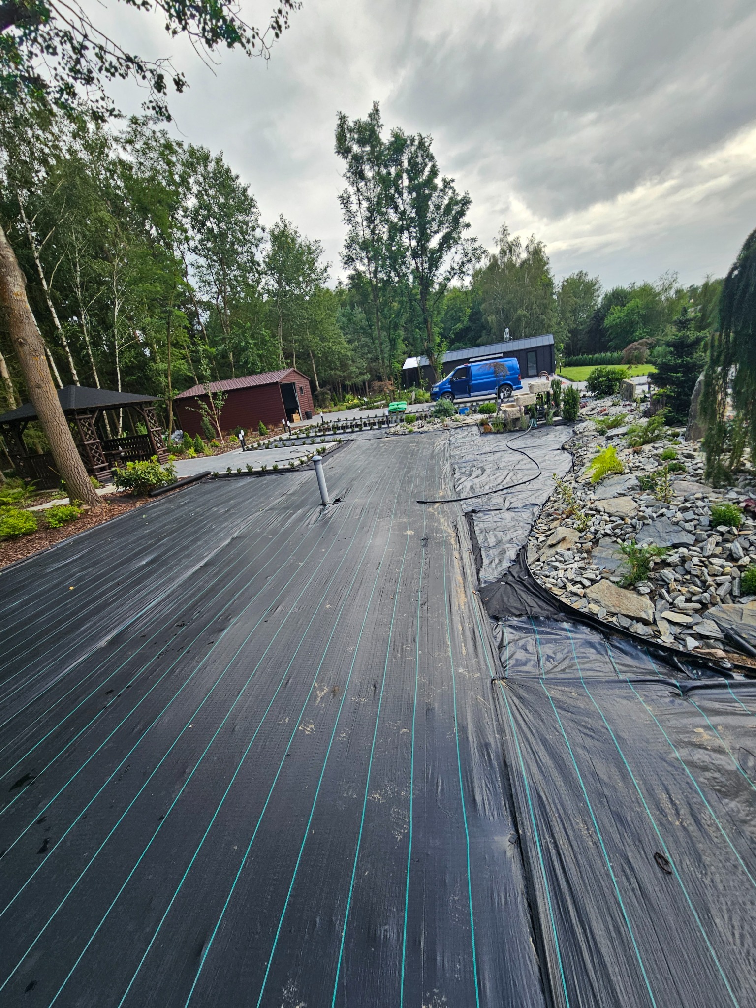 Rozłożona czarna agrotkanina z zielonymi paskami na przygotowanym terenie pod ogród, widoczne elementy przyszłego skalniaka z kamieniami, w tle altana, budynek gospodarczy i niebieski samochód...