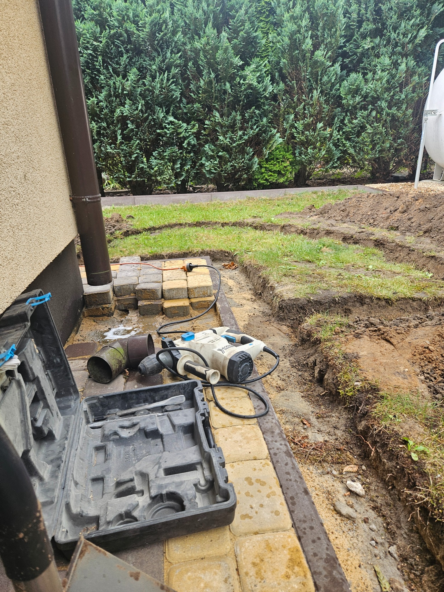 Przygotowanie terenu pod instalację nawadniania: wykopane rowki w ziemi obok ułożonej kostki, narzędzia pracy - wiertarka udarowa i otwarty kufer narzędziowy leżą na żółtej kostce brukowej, w tle...