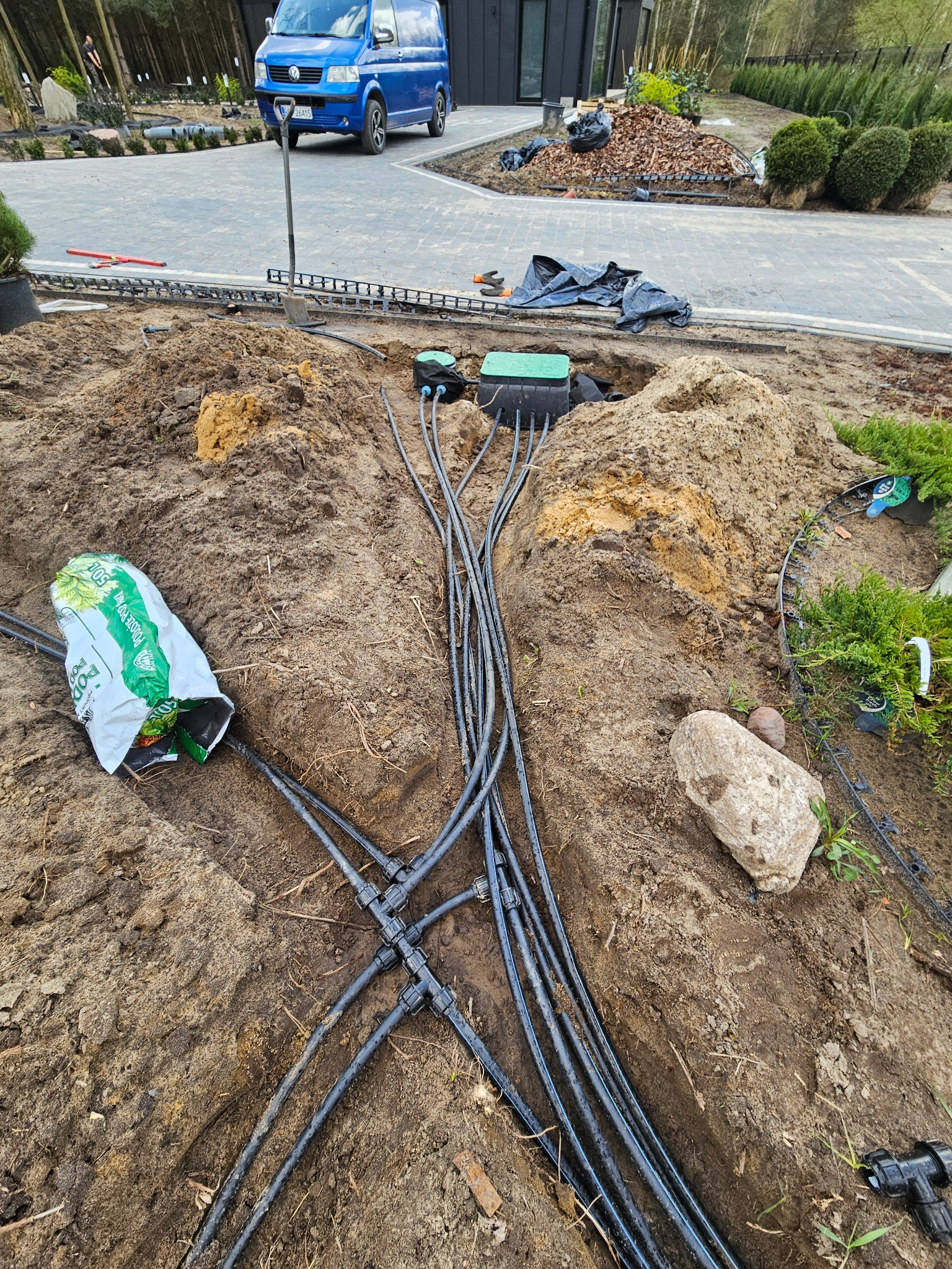 Rozbudowany system nawadniania w trakcie instalacji, widoczne czarne rury i złączki w wykopie, obok worek ziemi ogrodniczej i kamień, w tle ułożona kostka brukowa i zaparkowany niebieski bus.