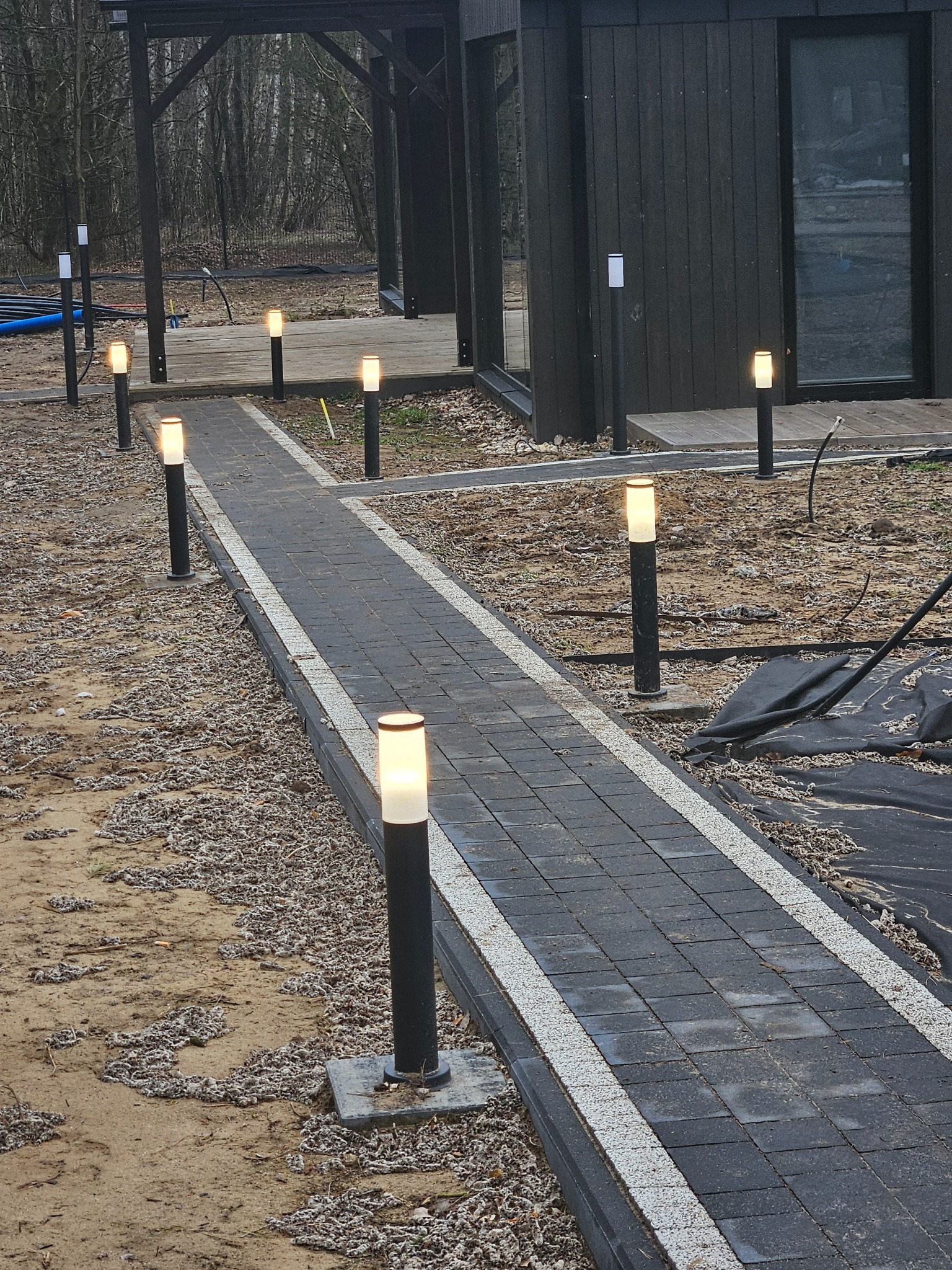 Ścieżka z ciemnej kostki brukowej obramowana jasnym kamieniem, oświetlona szeregiem nowoczesnych lamp ogrodowych, prowadząca do drewnianego budynku w tle.