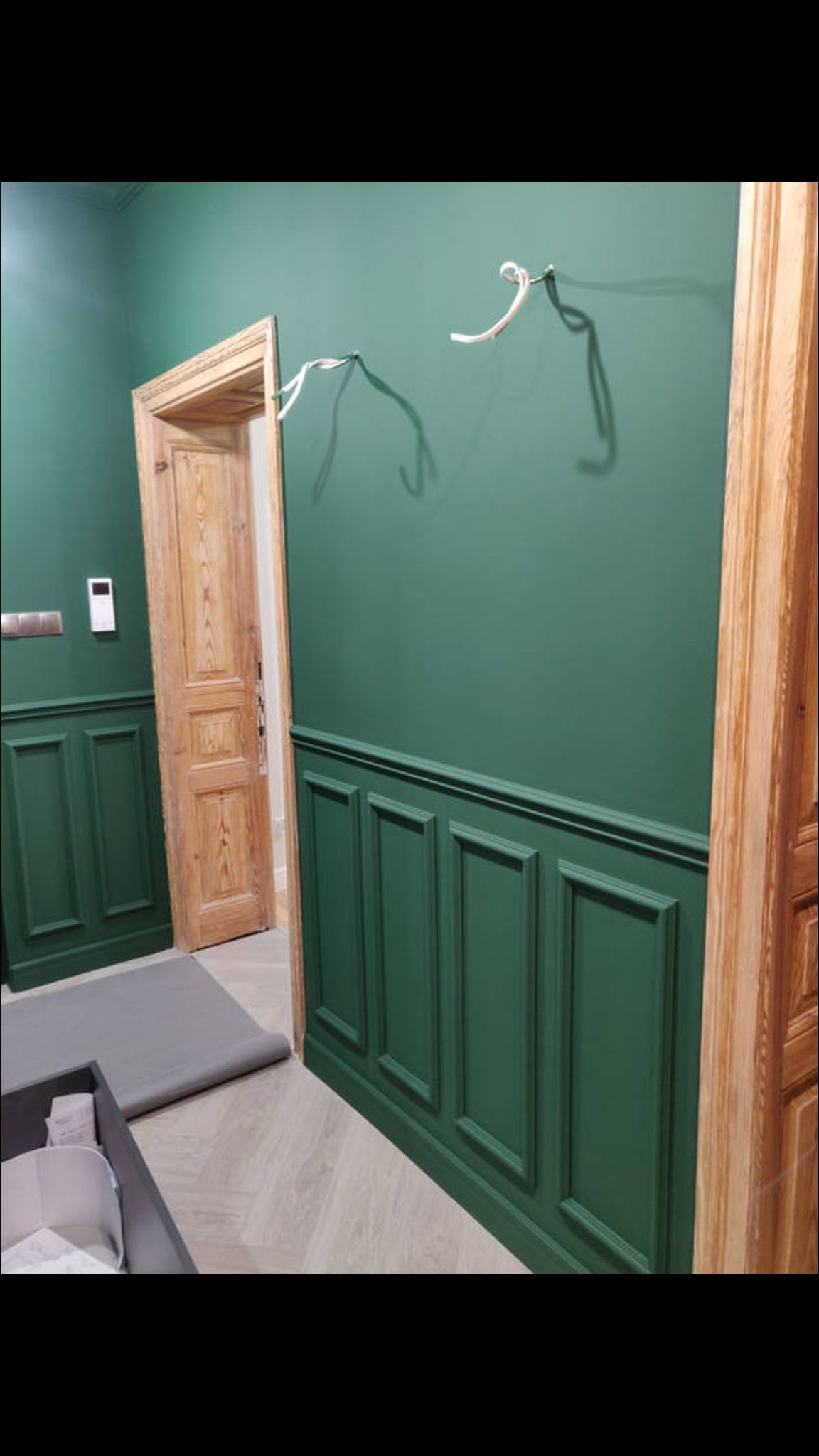Zielona ściana z drewnianą boazerią i otwartymi drzwiami z surowego drewna. Na ścianie widoczne ślady po instalacji.