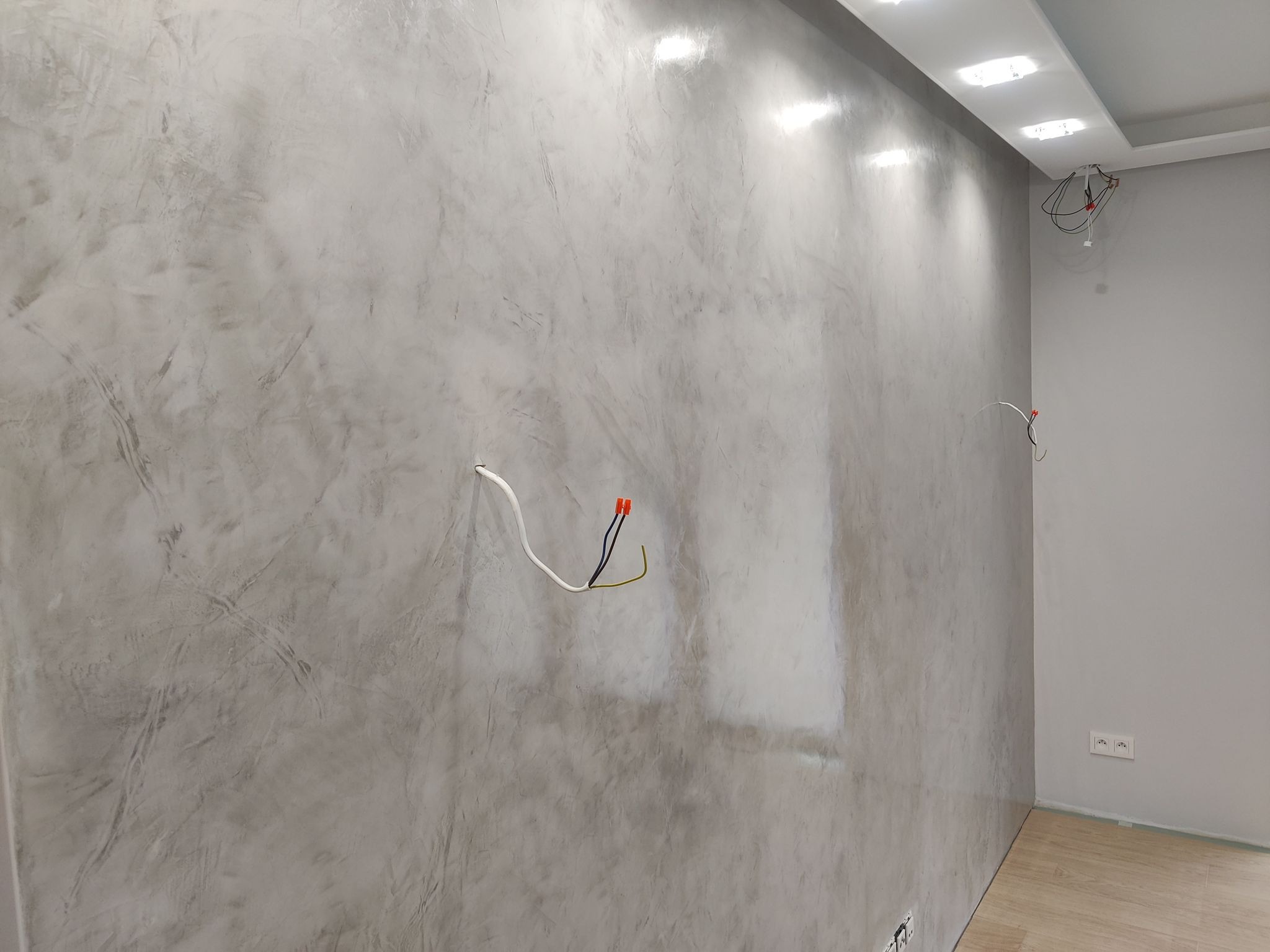 Ściana z efektem dekoracyjnym w odcieniach szarości, widoczne przewody elektryczne i gniazdka. Oświetlenie sufitowe w tle. Podłoga z paneli.