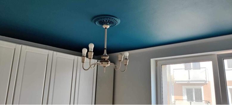 Błękitny sufit z dekoracyjną rozetą i żyrandolem w jasnym wnętrzu. Widoczne białe szafki i okno z widokiem na budynek.