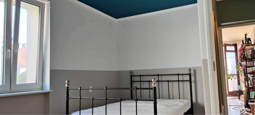 Pokój po remoncie: białe ściany z szarym pasem na dole, turkusowy sufit, czarne łóżko z białym materacem, okno i otwarte drzwi do innego pokoju.